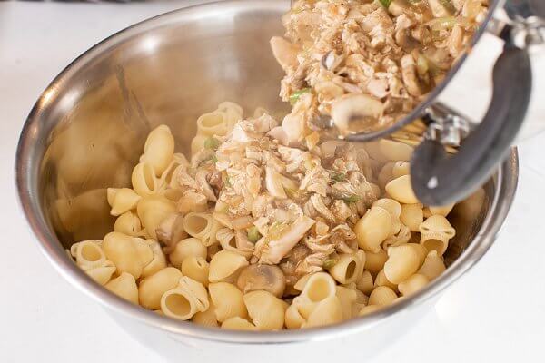Shredded turkey and mushroom mixture being added to cooked pasta in a metal bowl.