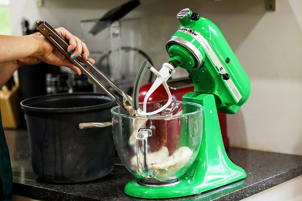 Adding cooked chicken breasts to a stand mixer with tongs.