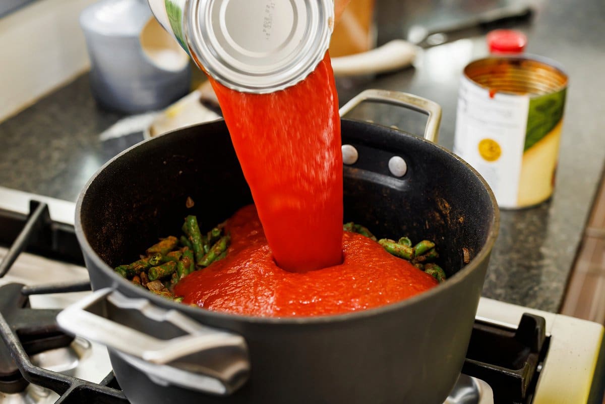 Pouring tomato sauce over green beans in large pot.