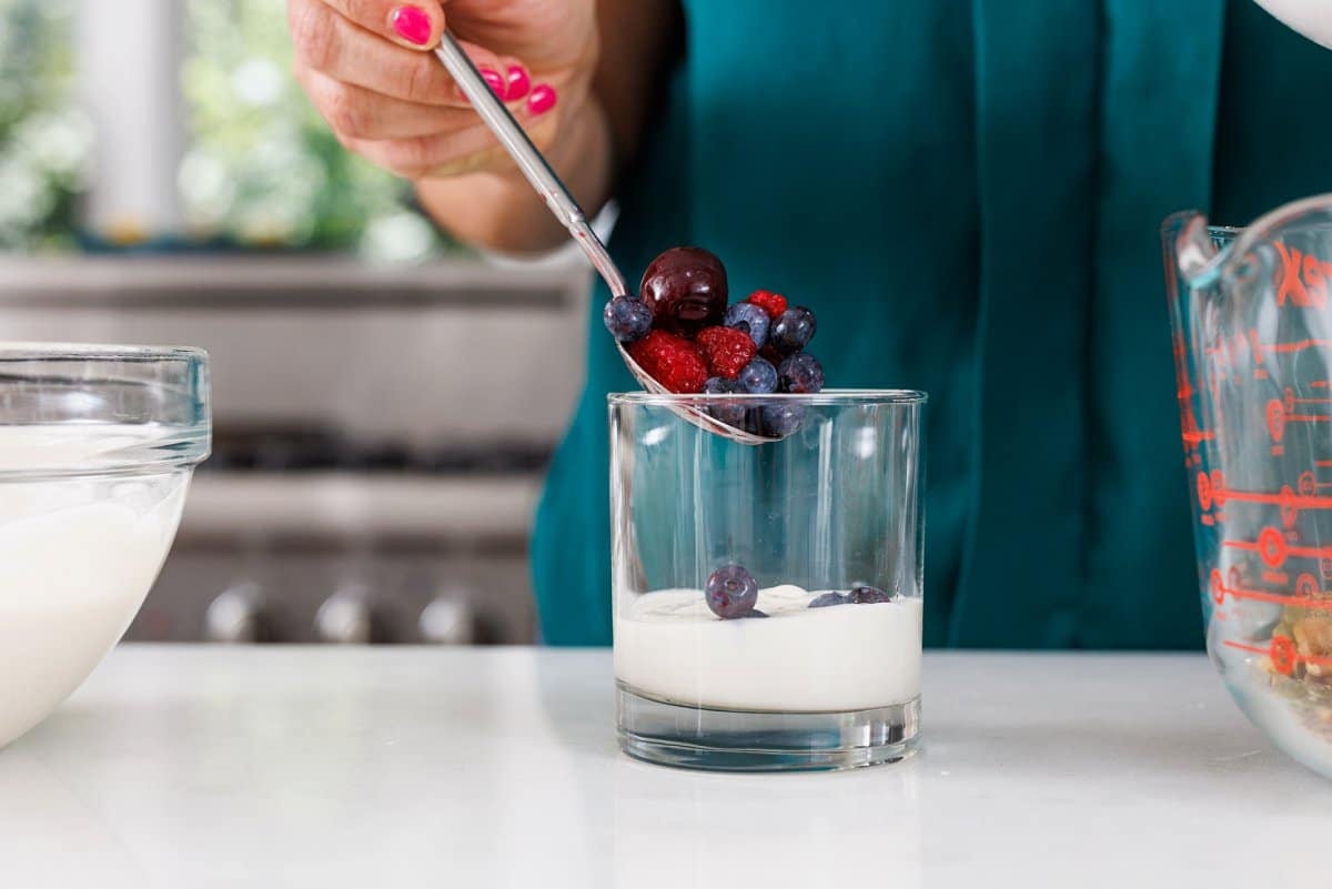 Adding mixed berries on top of yogurt in glass cup.