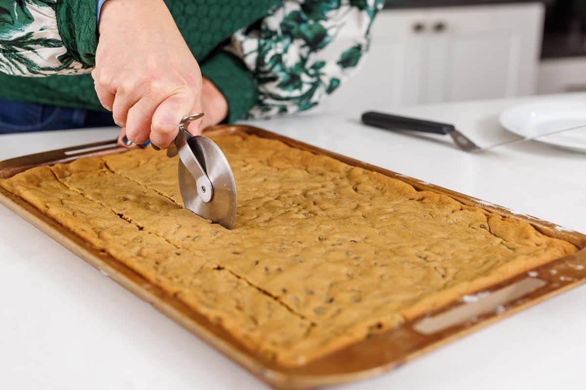 Using a pizza cutter to cut baked hermit bars into squares.