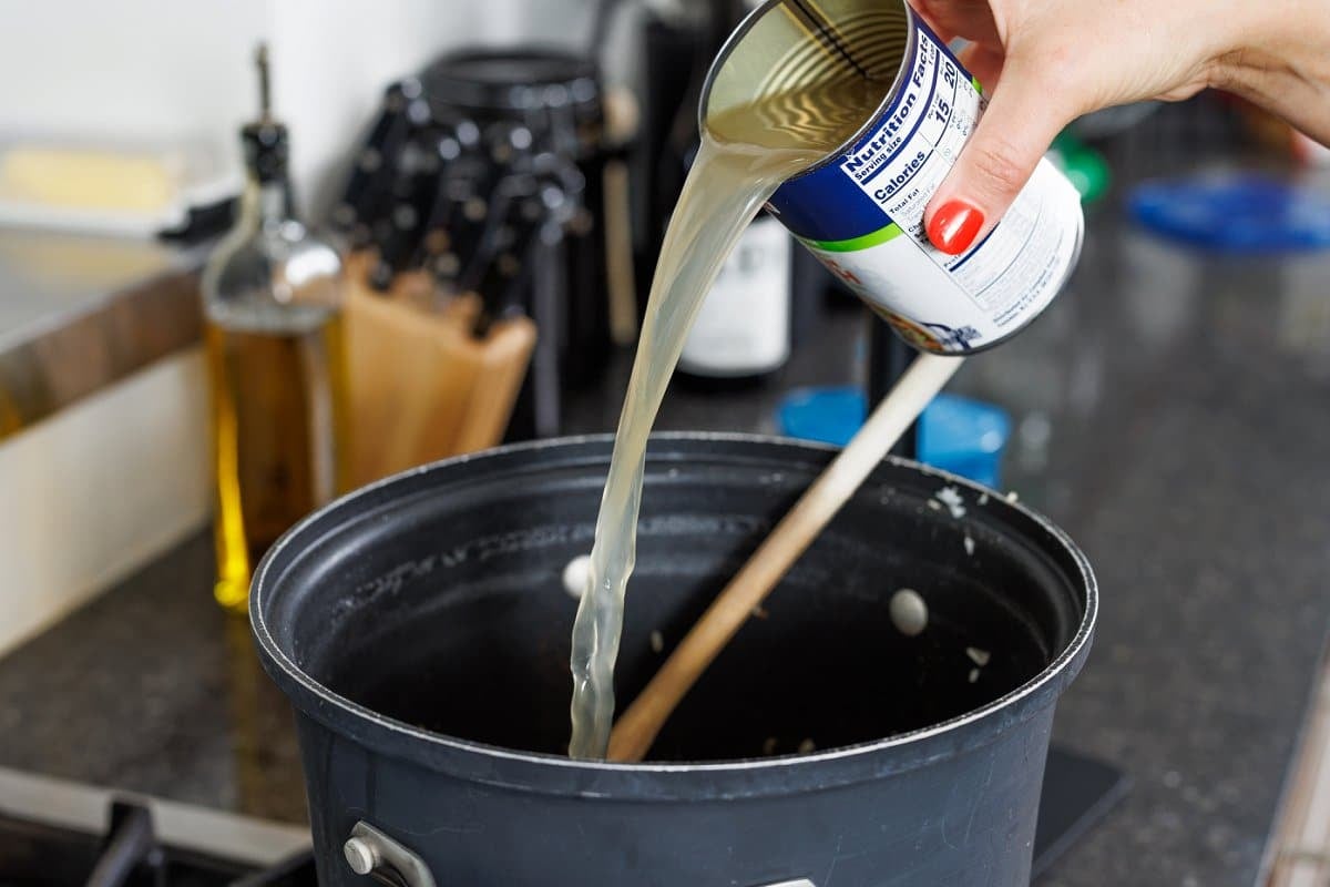 Adding chicken broth out of a can to the pot of rice.