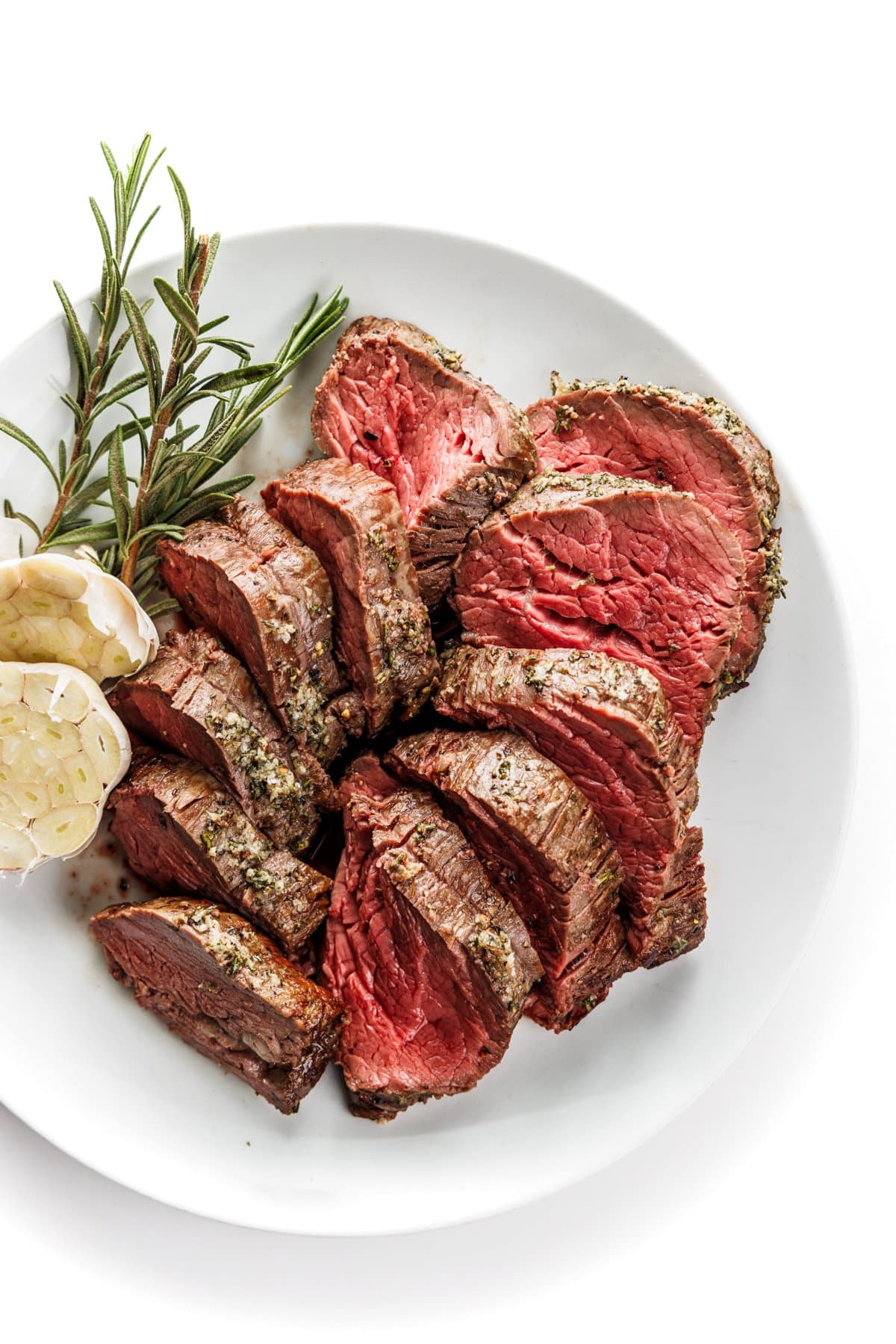 Garlic rosemary beef tenderloin on a white plate next to rosemary strands and garlic cloves.