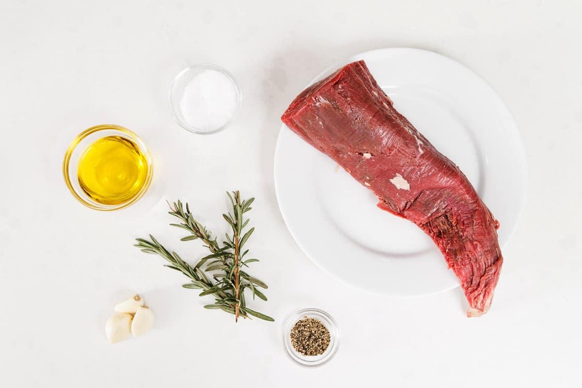 individual ingredients for rosemary beef tenderloin arrayed against a white surface.