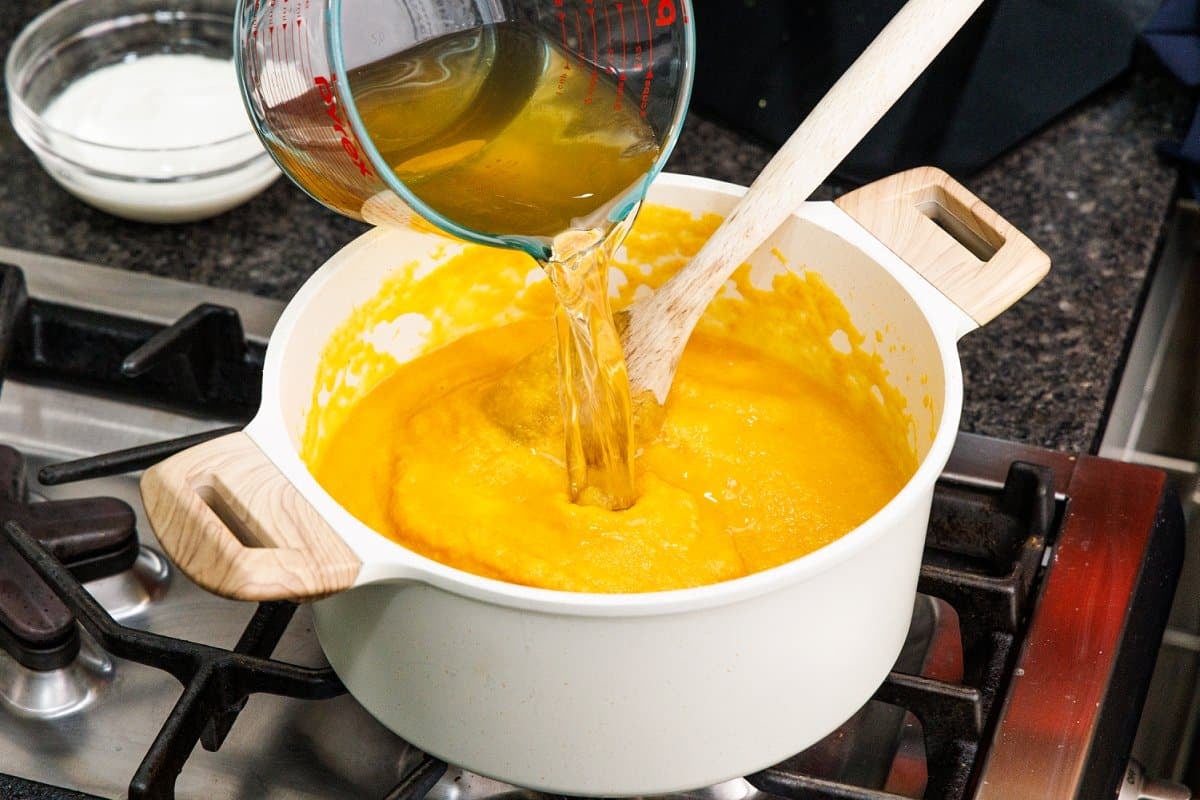 Chick broth being added to vegetable soup in a white pot on the stove.