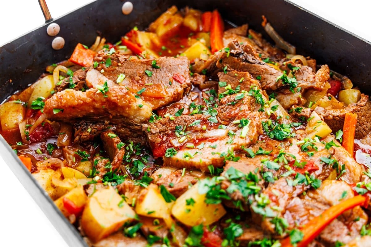 Roasting pan full of cooked beef brisket, potatoes, carrots, and garnished with fresh herbs.