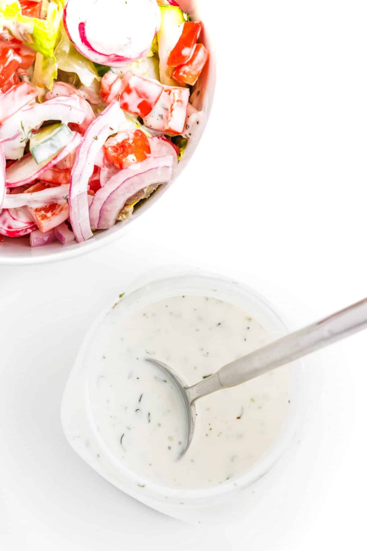 Top view of 5-ingredient dill ranch dressing in a jar with a spoon, next to a green salad topped with the dressing.