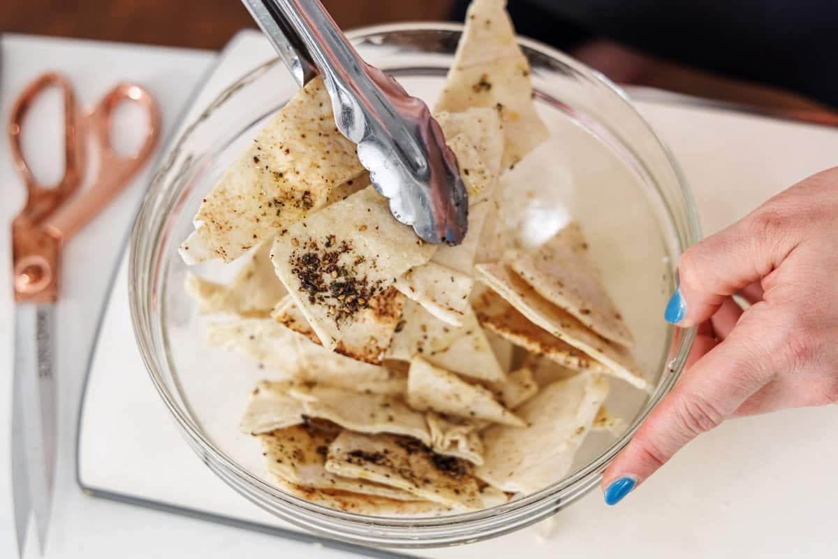 Seasoning pita bread triangles in oil and za'atar in a glass bowl.