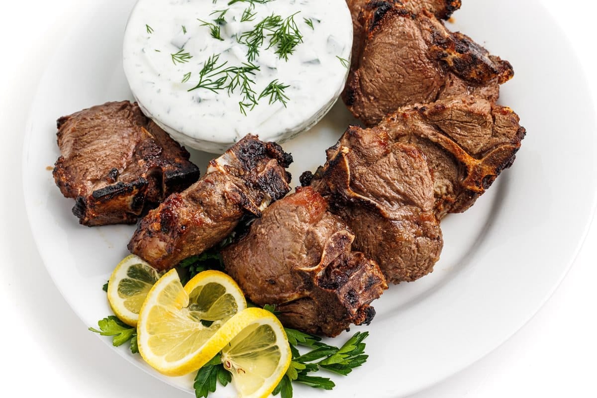 Lamb chops on a plate with tzatziki sauce in a small bowl next to them, garnished with lemon slices.