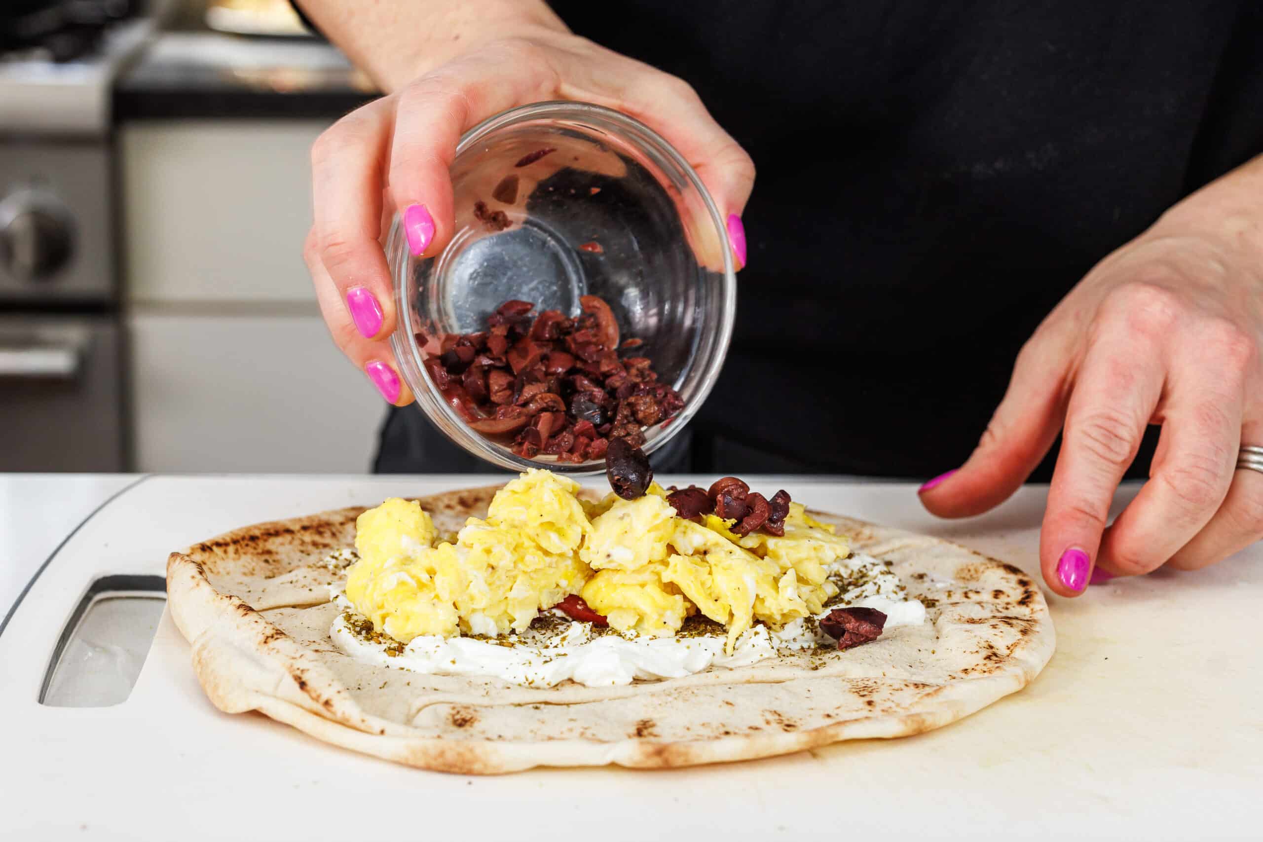 Pouring halved olives on breakfast pita.