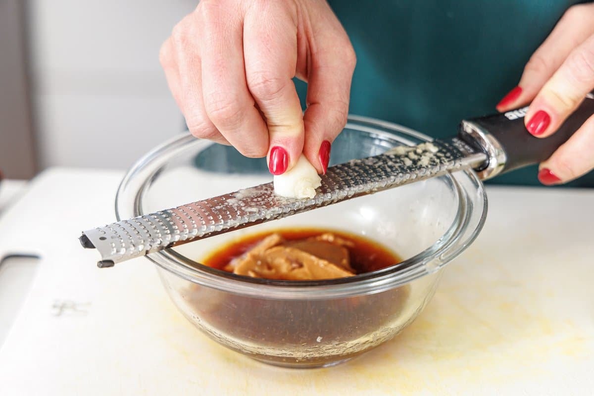Grating garlic clove into glass bowl with soy sauce and peanut butter.