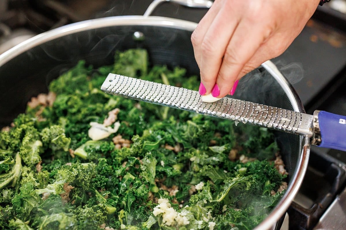 Grating garlic into a large skillet of kale and sausage.
