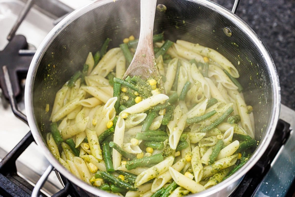 Stirring cooked pasta and veggies in pesto in a pot.