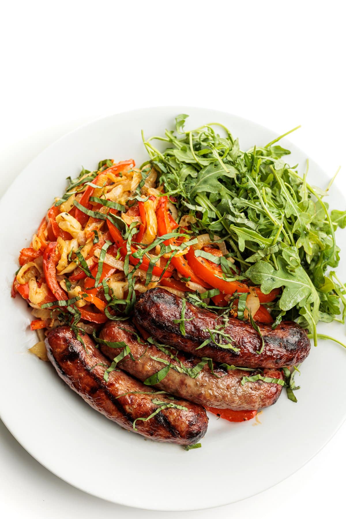 Grilled bratwursts with grilled peppers and onion on a plate with arugula and garnished with basil ribbons.