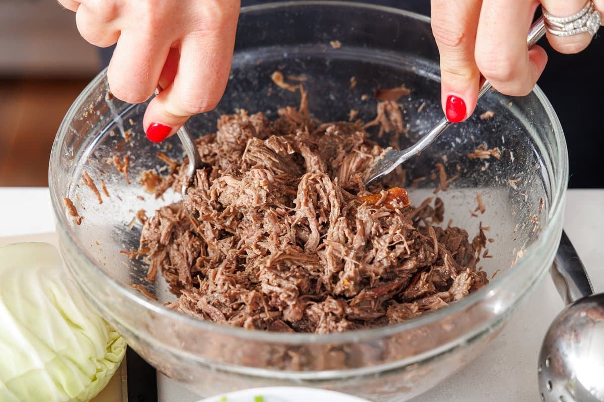 Liz shredding beef in a glass bowl with two forks.
