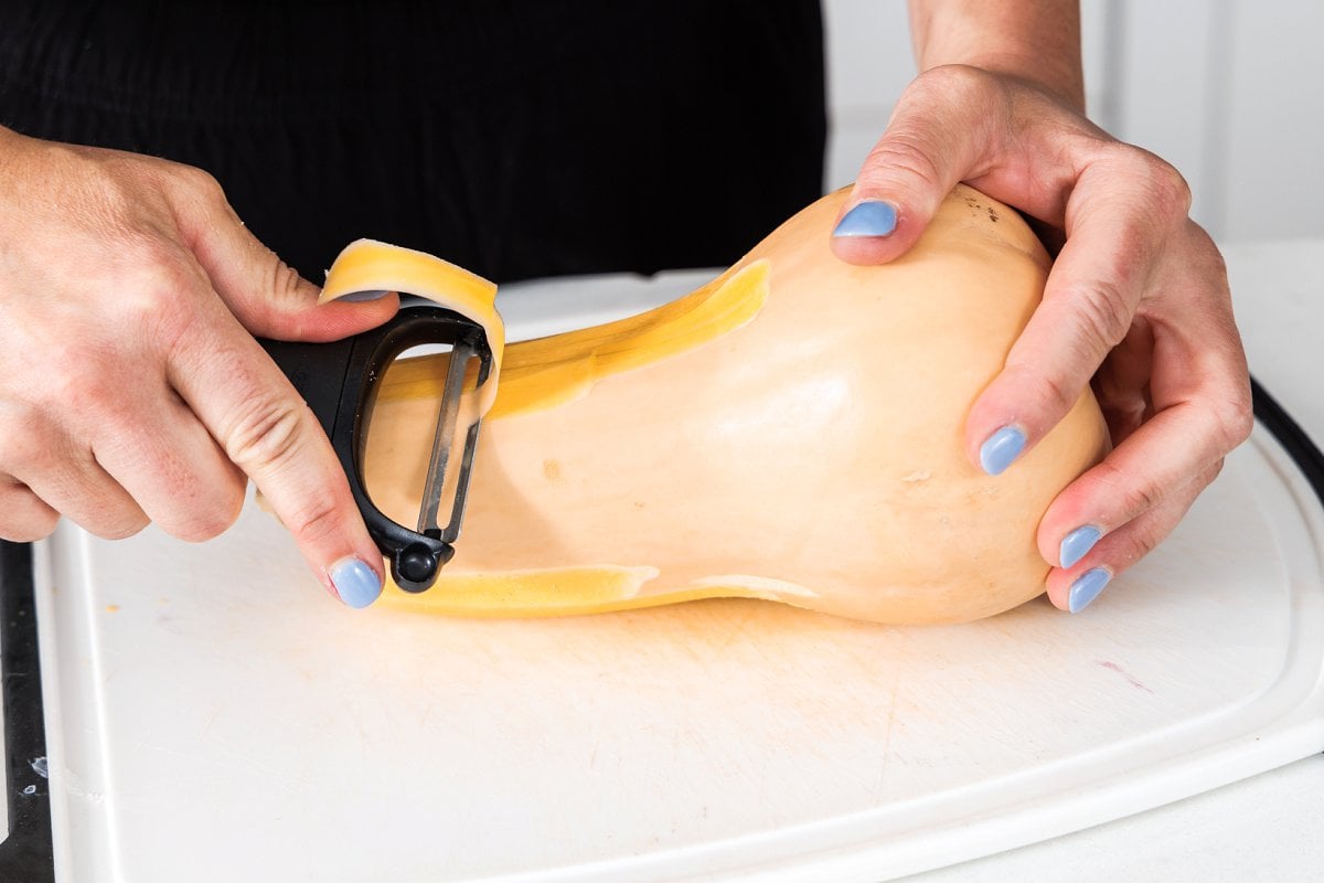 Using a vegetable peeler to peel whole butternut squash.