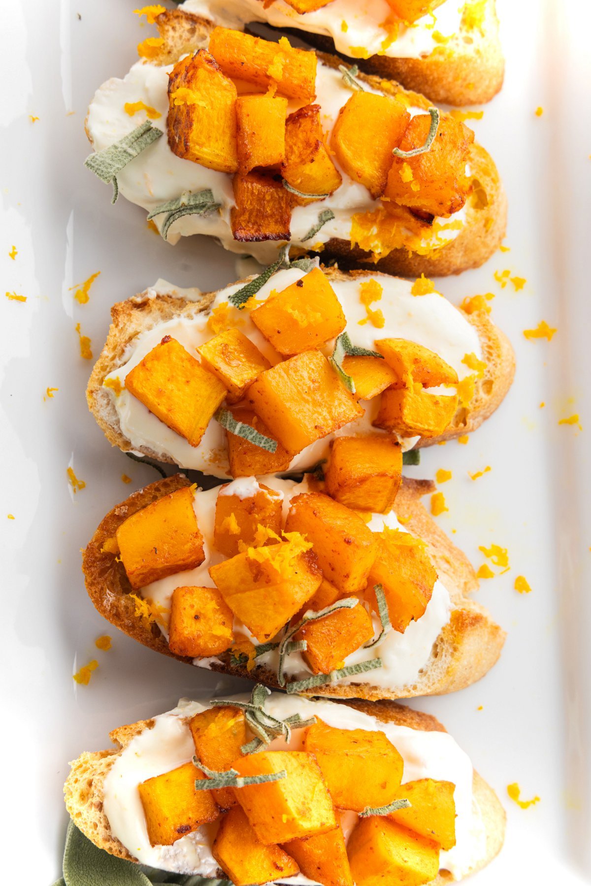 Four butternut squash crostini with whipped goat cheese and sage on a white plate and garnished with orange zest.