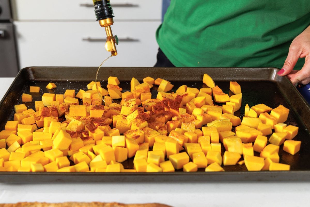 Drizzling olive oil over cut up butternut squash on a baking sheet.